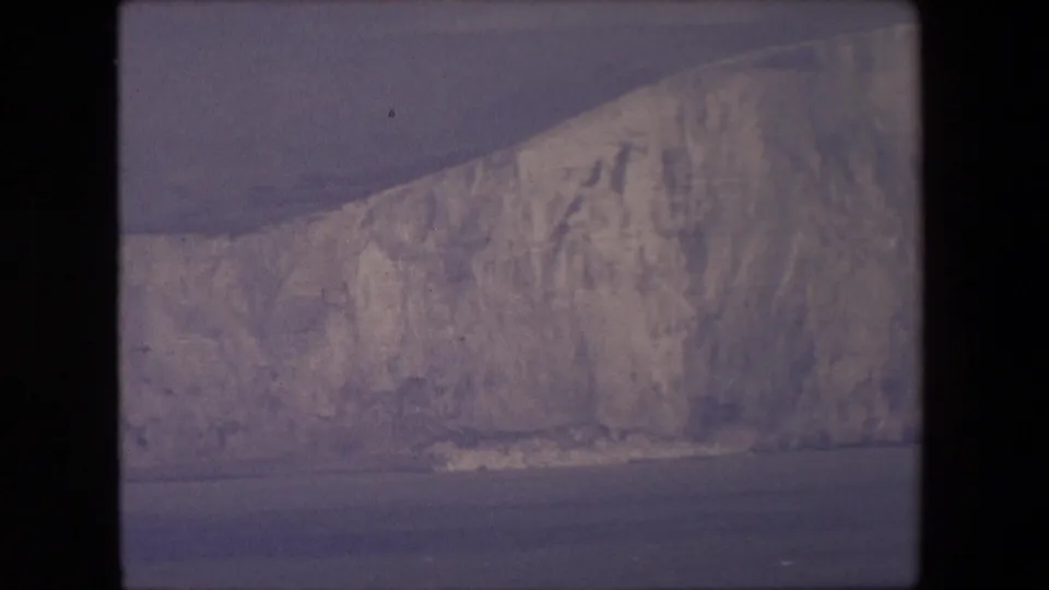 1967 8mm home-movie frame showing a closer pale cliff wall with vertical weathering marks above dark blue-gray sea.