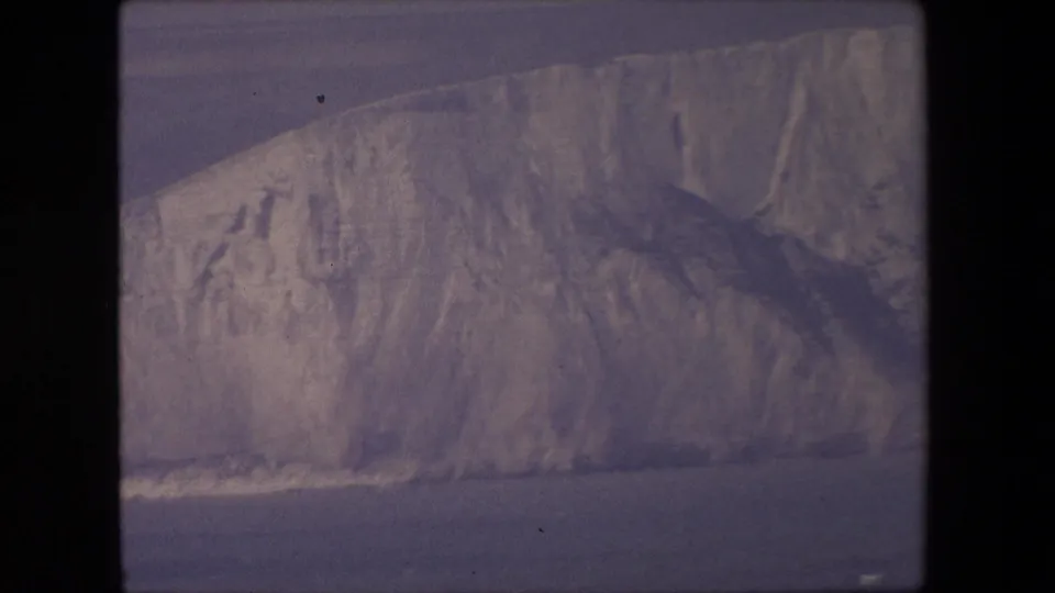 1967 8mm frame showing a tall, textured pale sea cliff dominating the view, with water at its base and a hazy sky above.