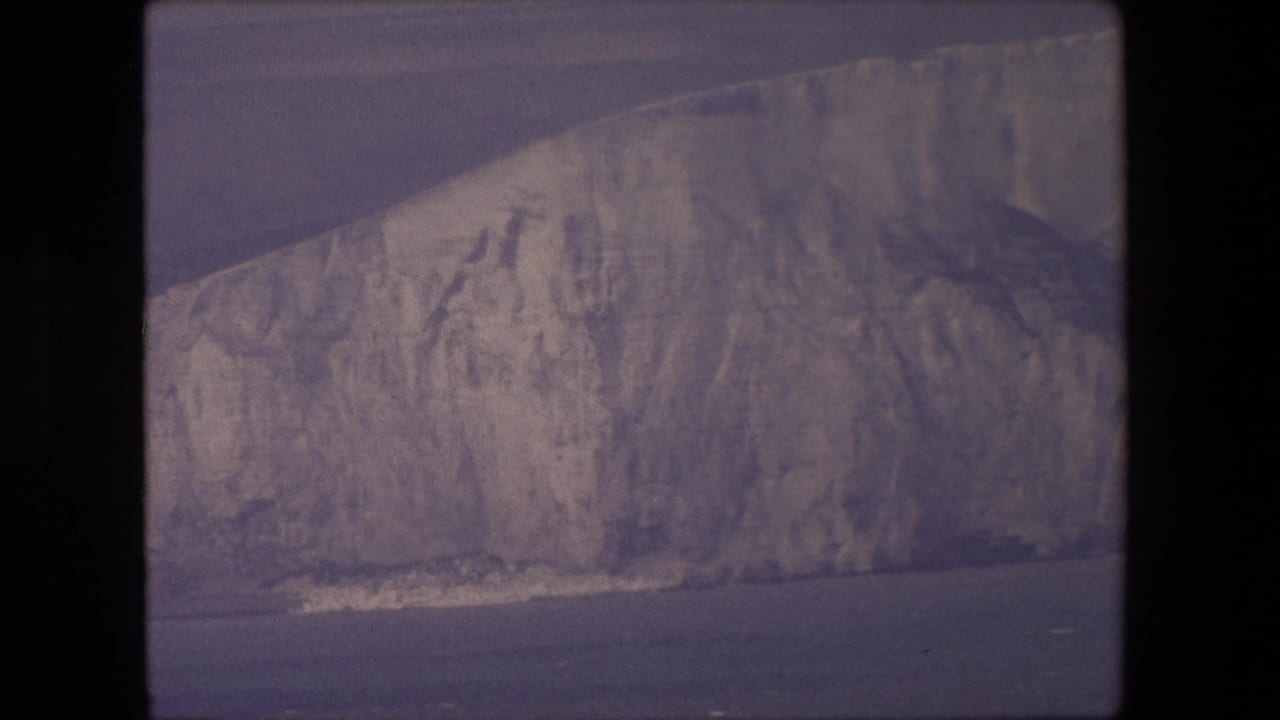 1967 8mm home-movie frame of a pale coastal cliff above open water, with a narrow wave line along the base and soft purple-gray film color.