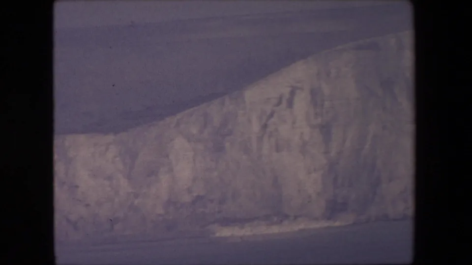 1967 8mm home-movie frame showing a broad chalk cliff wall across the sea, with muted light, visible grain, and surf along the lower edge.