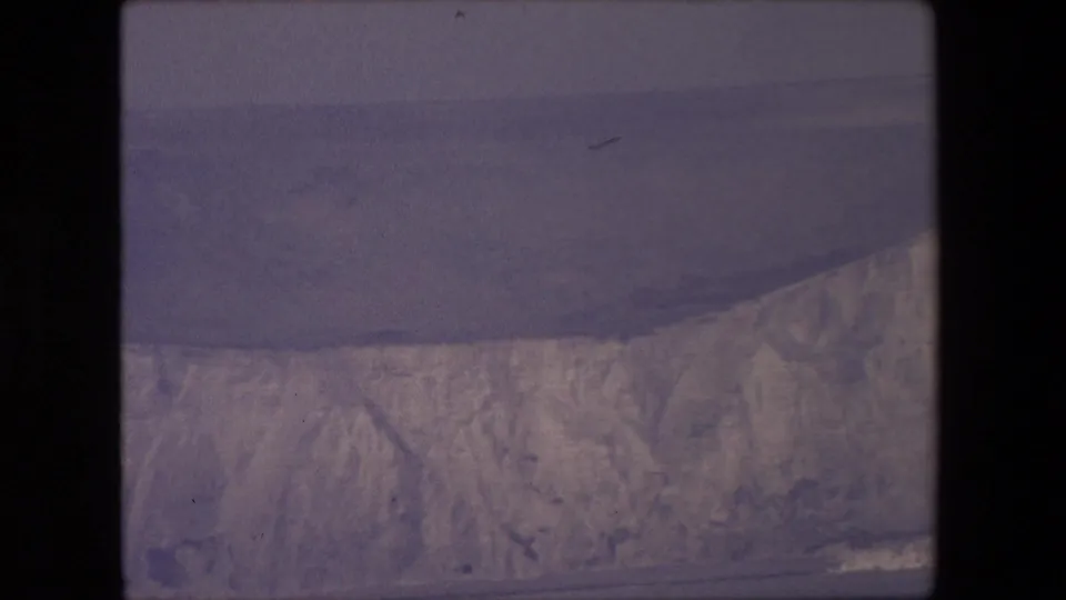 1967 8mm home-movie frame of a pale cliff edge and open sea, with soft focus, visible film grain, and subdued purple-gray tones.