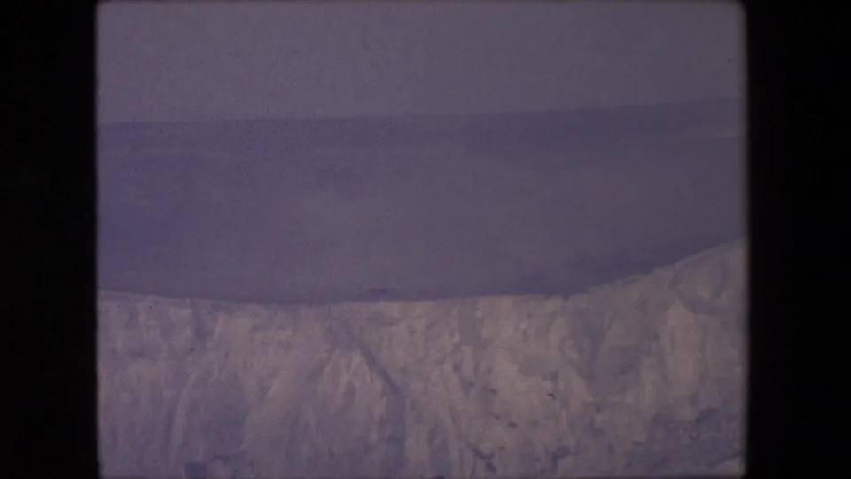 1967 8mm home-movie frame showing pale coastal cliffs and a dark sea beneath a hazy overcast sky, with no people or structures visible.