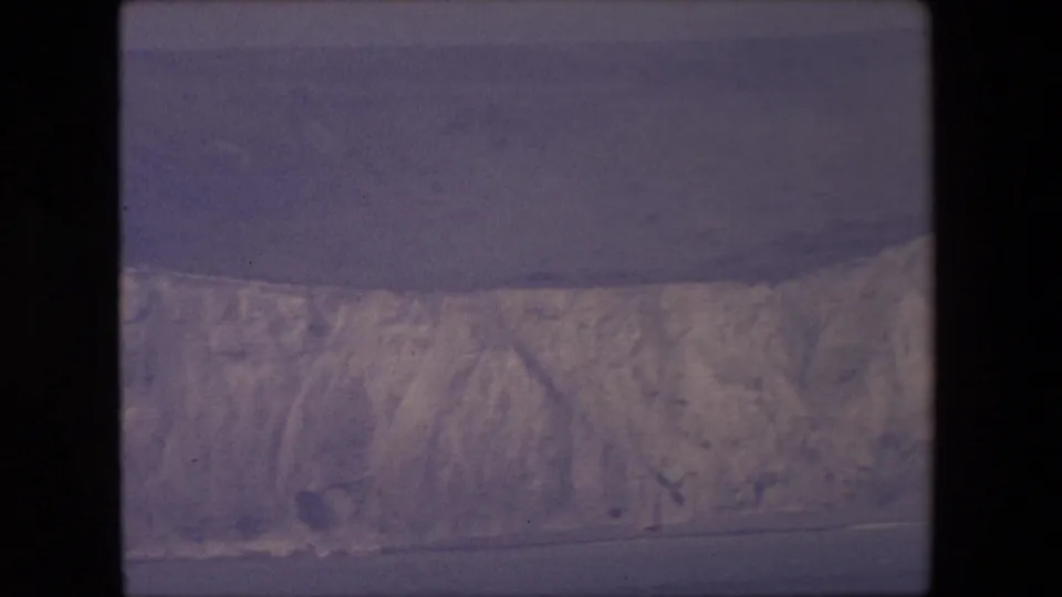 1967 8mm frame of a pale cliff line overlooking dark water, with subdued color, soft focus, and a hazy horizon.