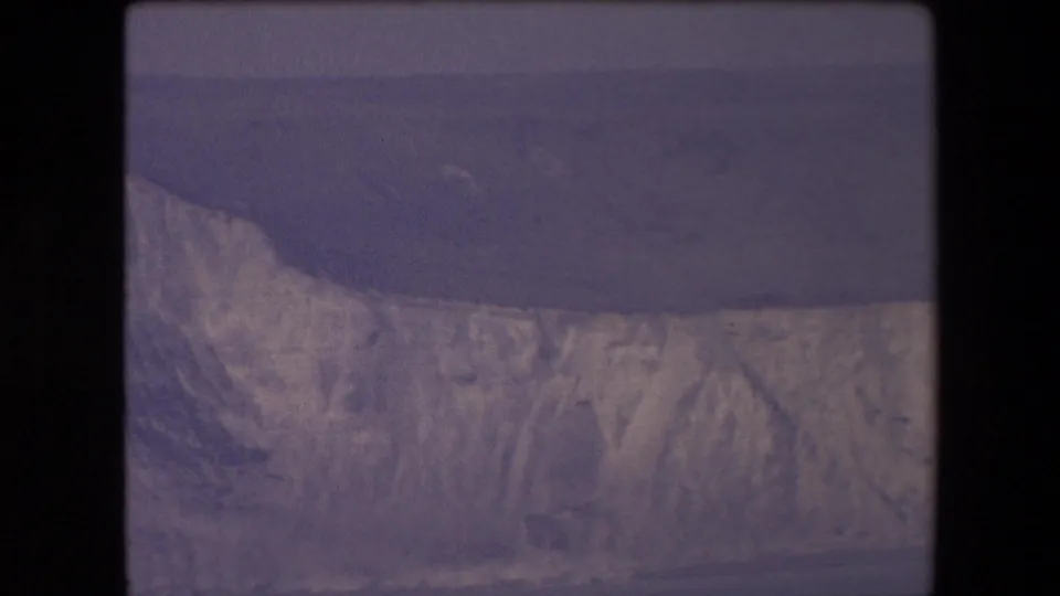 1967 home-movie frame showing chalky coastal cliffs, dark sea, and a broad shadowed headland or slope in soft 8mm color.