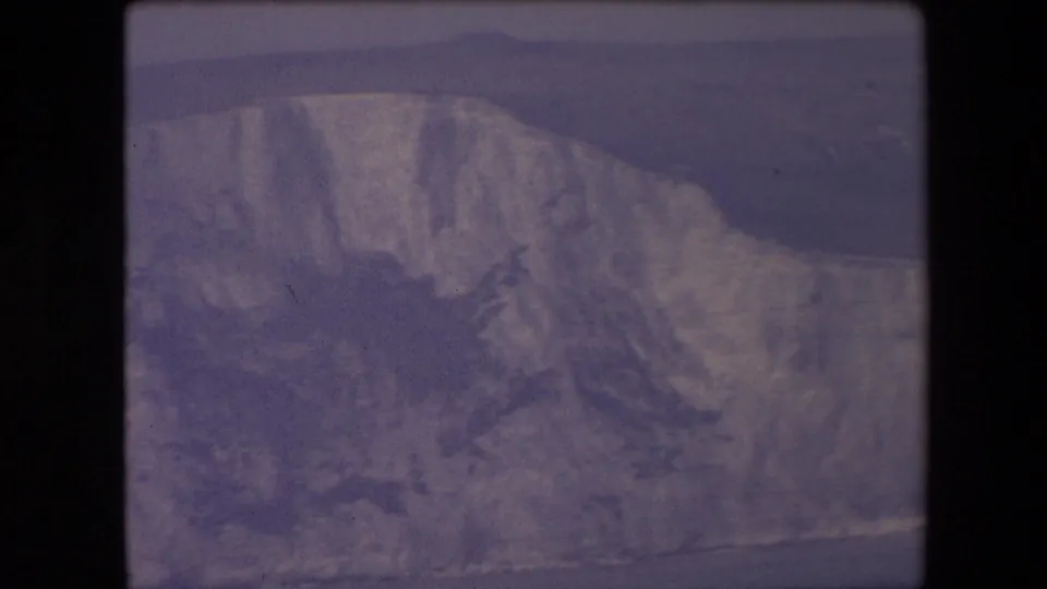 1967 8mm frame of a rugged pale cliff face beside dark sea, rendered with visible grain, muted color, and soft handheld framing.