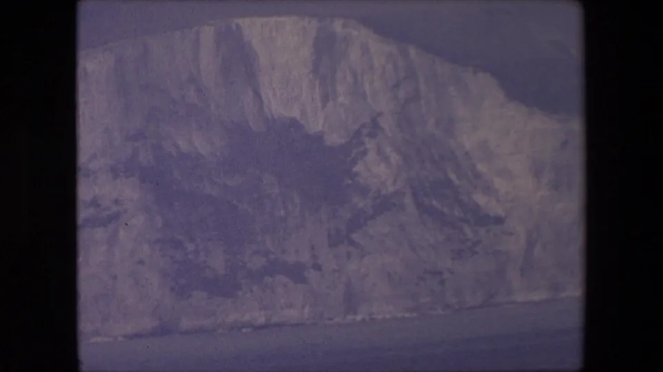1967 archival 8mm frame showing pale chalk cliffs along a coastline, with a dark sea band at the base and a muted overcast sky above.