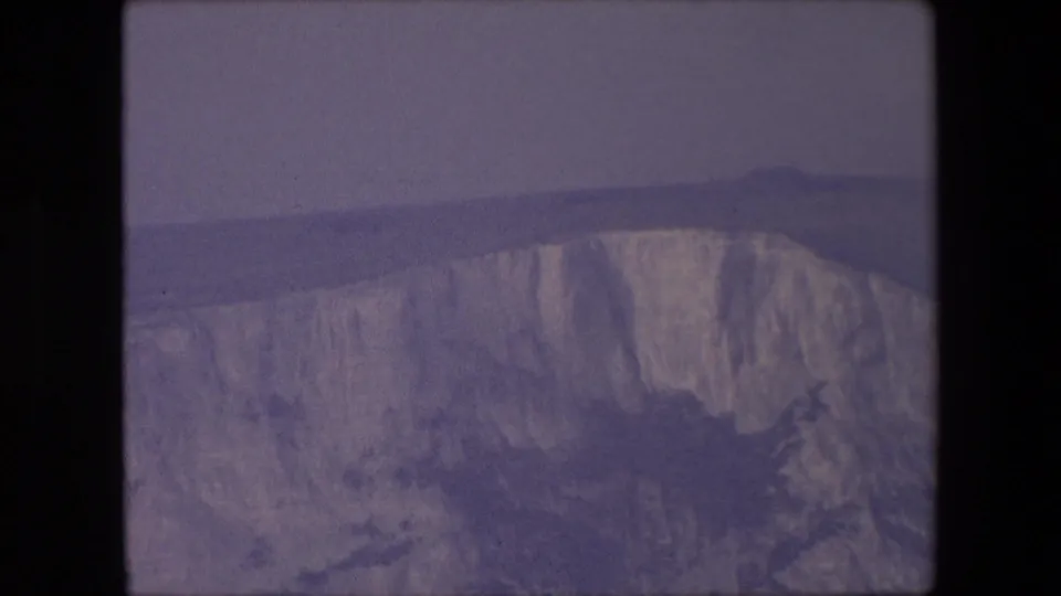 1967 archival 8mm frame of a broad chalk cliff face seen from a distance, with a subdued sky and a flat horizon line behind it.