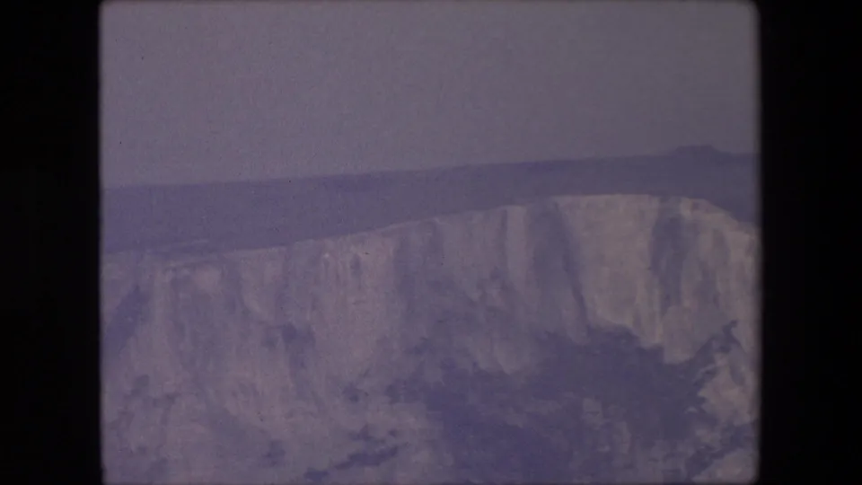 1967 archival 8mm frame showing chalk cliffs from a distant viewpoint, with the sea and horizon softened by haze and film exposure.