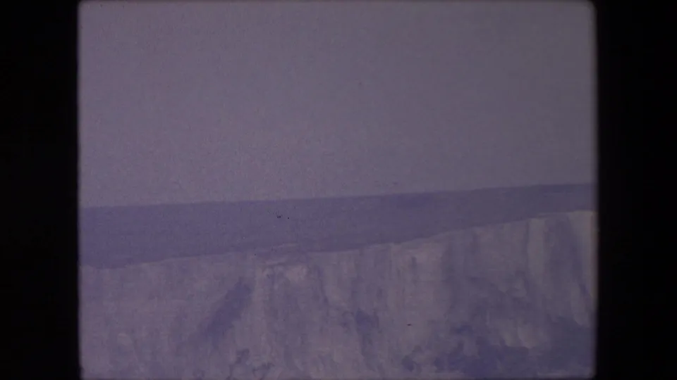 1967 archival 8mm frame of a distant chalk cliff ridge under a muted sky, with a dark horizontal band suggesting sea or far shoreline.