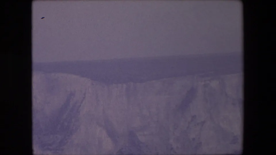 A small seabird appears near the upper left of a faded 8mm coastal view, with pale cliffs and dark sea below in the stated England footage.