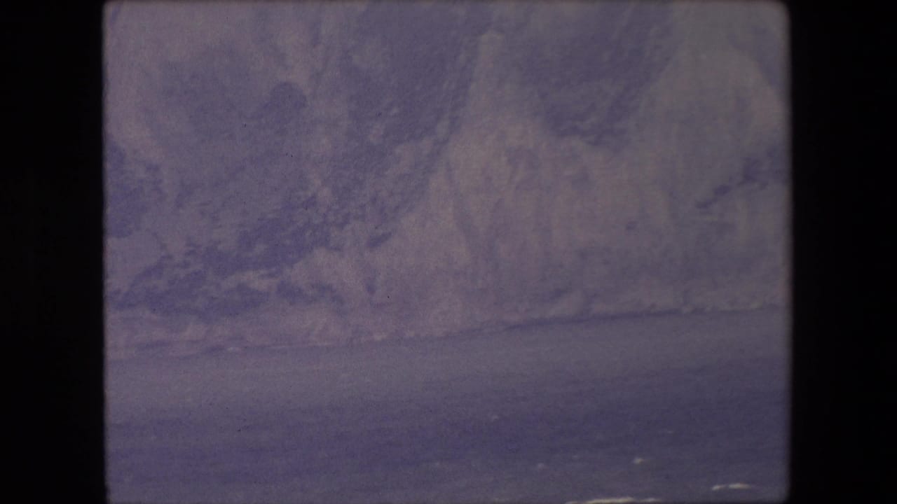 Soft, purple-tinted view of pale sea cliffs above dark water.
