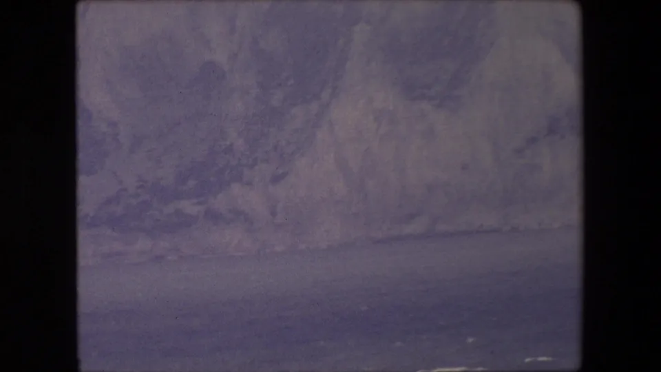 1960s 8mm frame showing pale coastal cliffs and dark water under a muted sky, with heavy film border visible.