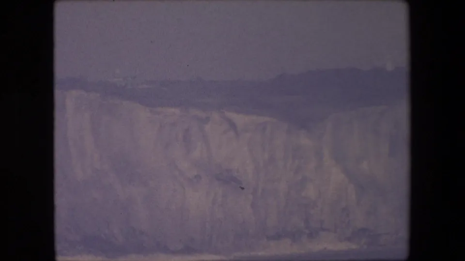 Archival 8mm coastal view with a pale cliff line, dark distant headland, and subdued overcast sky.