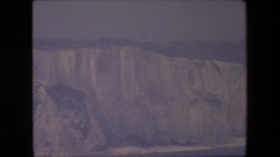 1960s home-movie frame showing a long pale cliff face beside dark sea, rendered with soft focus and purple-blue color shift.