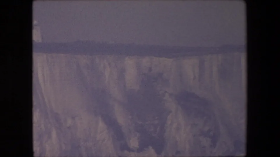 Archival coastal frame showing pale cliffs, dark water, hazy horizon, and a darker landform or shadowed cliff area near the lower right.