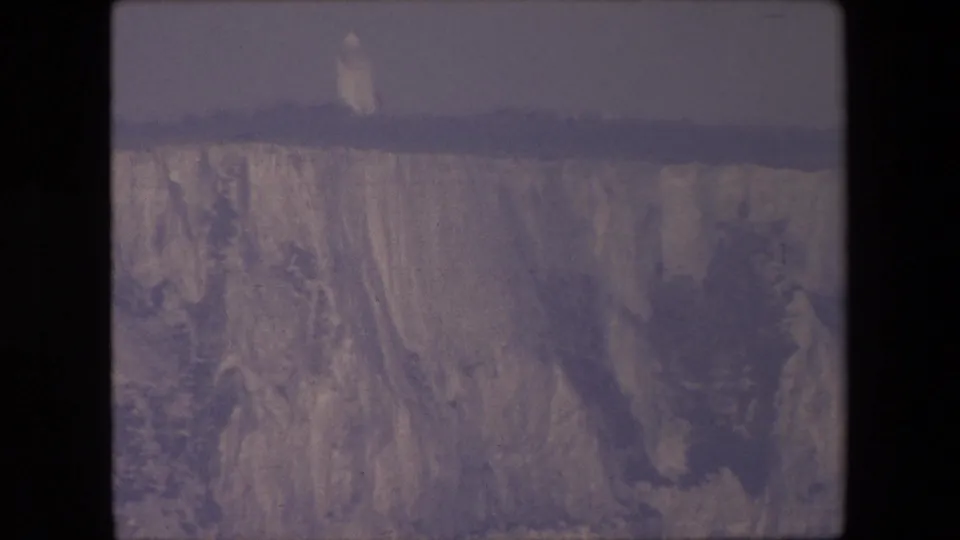 1967 8mm home-movie frame showing a pale, streaked cliff face under an overcast sky, with a small distant tower-like form near the upper edge.