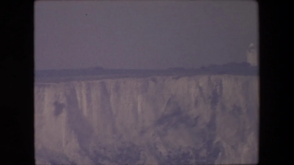 1967 8mm frame of a pale coastal cliff under flat gray light, with a small lighthouse-like or tower-like structure visible near the right edge.
