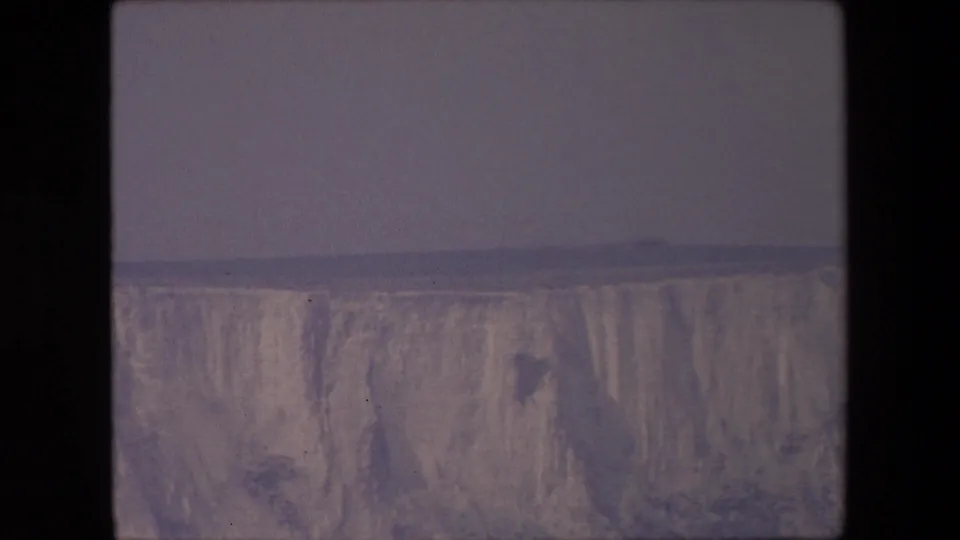 1967 8mm home-movie frame holding on the lower portion of a pale, streaked cliff face beneath a muted gray-blue sky.