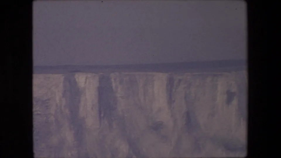 1967 8mm frame showing the cliff face from a distance, with a flat horizon line, gray overcast sky, and softly faded color.