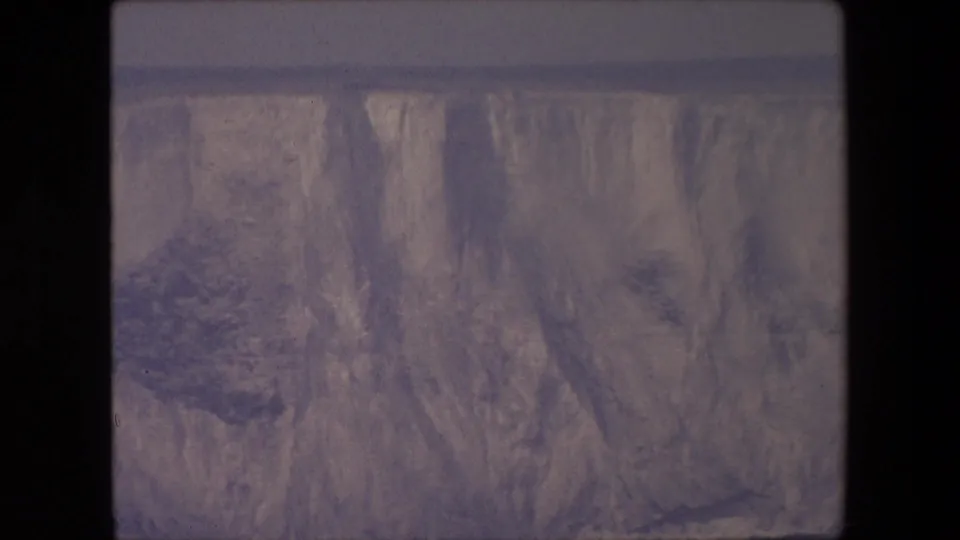 1967 metadata-supported London 8mm frame showing a steep eroded rock or cliff face with sparse dark vegetation and faded gray-purple color.