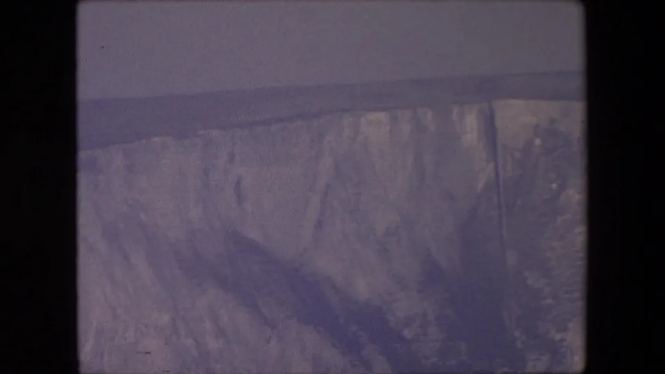 A faded 8mm frame shows a steep, textured glacier face rising above water, with no people or structures visible.