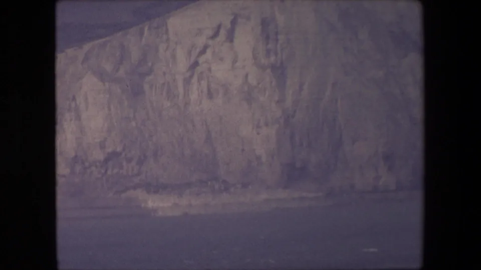 The frame presents a closer view of the glacier's rough vertical face, pale ice striations, and a narrow strip of water at the base.