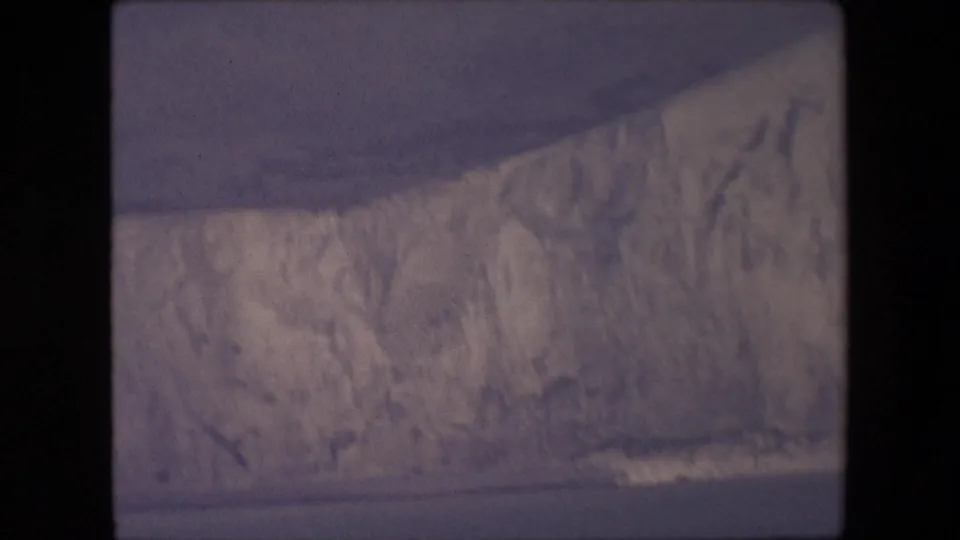 A faded archival frame shows a massive glacier front with a dark sloping shadow across the ice and water visible along the bottom edge.