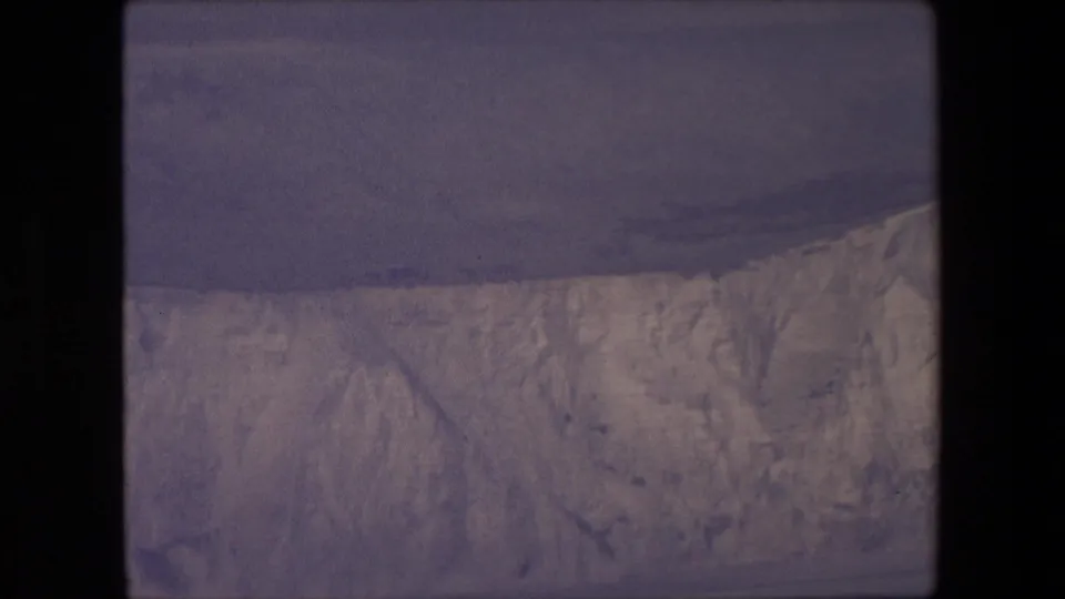 1967 8mm frame with a broad pale ice or chalk-like cliff face above a dark horizontal band of water, recorded in soft handheld home-movie style.