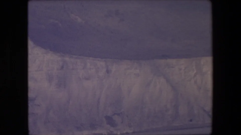 1967 8mm frame showing a lower section of pale ice cliffs and a wide dark water band, with visible film grain and subdued color.