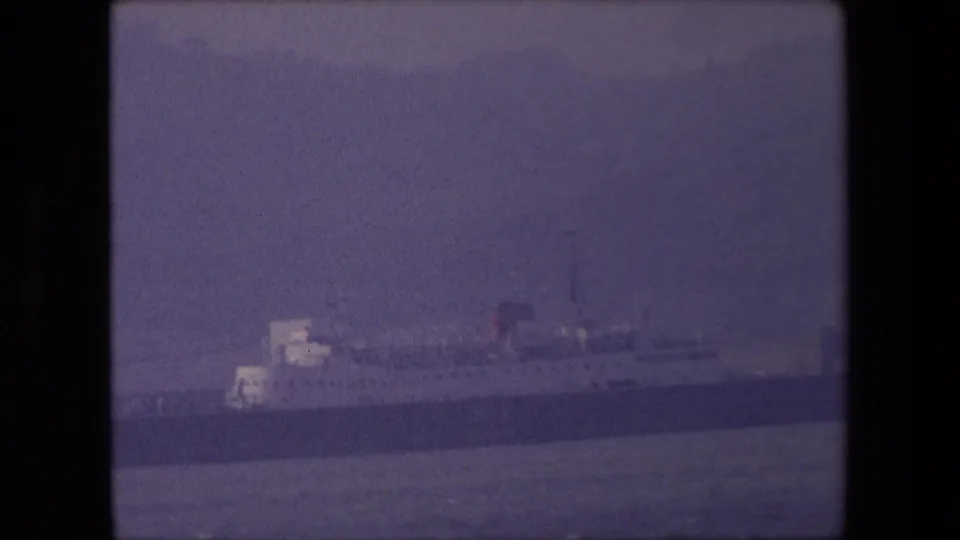 1967 8mm frame showing a distant large ship on dark water with pale ice cliffs behind it, filmed in muted color with no readable markings.