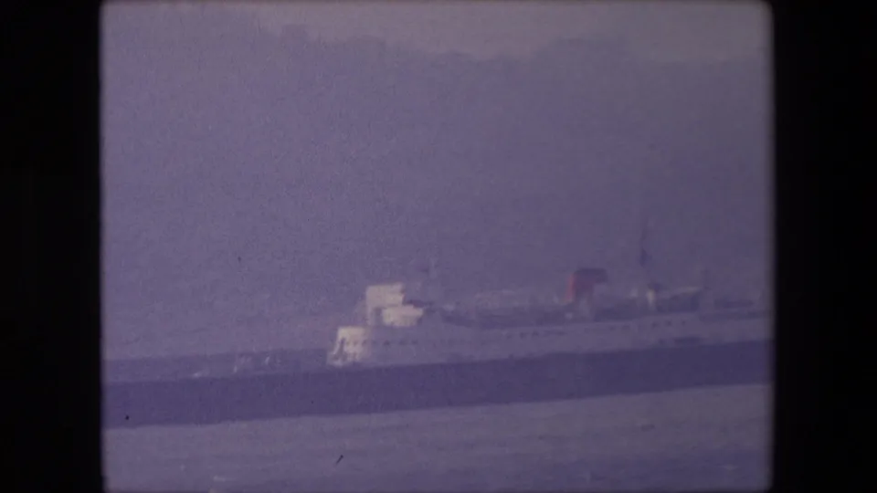 1967 London 8mm frame showing a distant large passenger ship with a white upper structure and red funnel across calm water under a hazy sky.