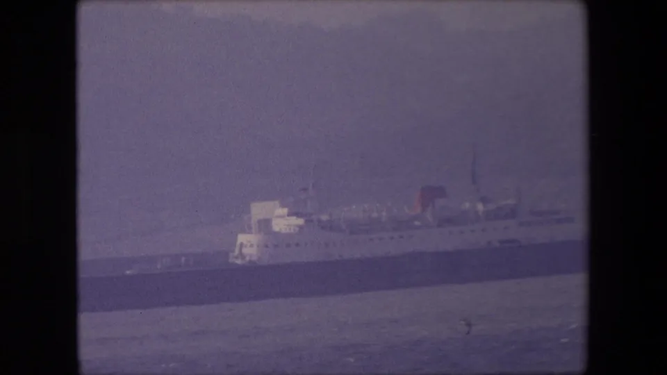 1967 London 8mm still of a large passenger vessel lying along the harbor, with calm water in front and a muted overcast atmosphere.