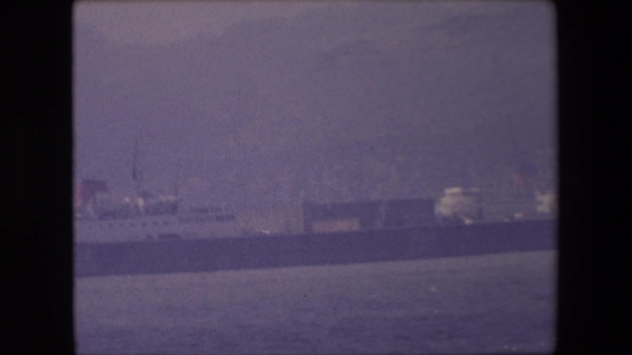 Distant long ship on calm water in London-associated 1967 8mm footage, seen through soft haze with shoreline structures behind it.