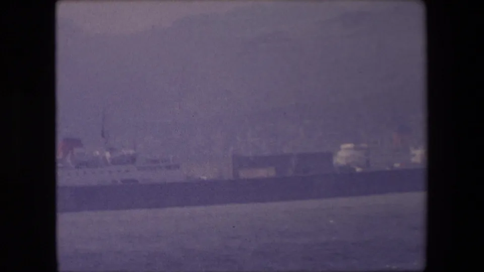 Distant long ship on calm water in London-associated 1967 8mm footage, seen through soft haze with shoreline structures behind it.