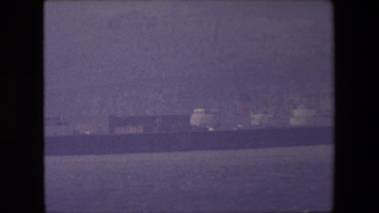 Long ship seen farther across rippled water with low buildings behind it.