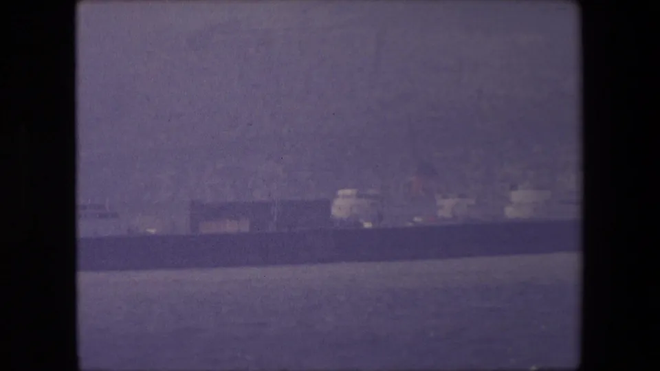 A long vessel continues across calm water in the distance, with faint shoreline buildings and the purple-tinted softness of 8mm film.