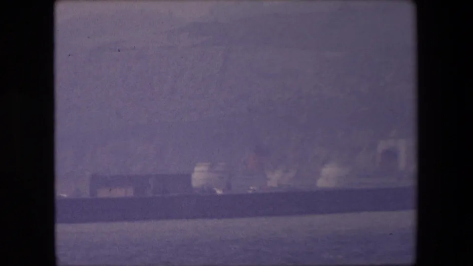 Soft 1967 London-associated 8mm frame showing the distant ship low in the composition, with calm water and indistinct waterfront structures.