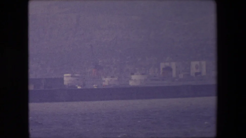 The ship appears near a low shoreline with pale buildings or vessel structures behind it, recorded in grainy purple-tinted 8mm footage.