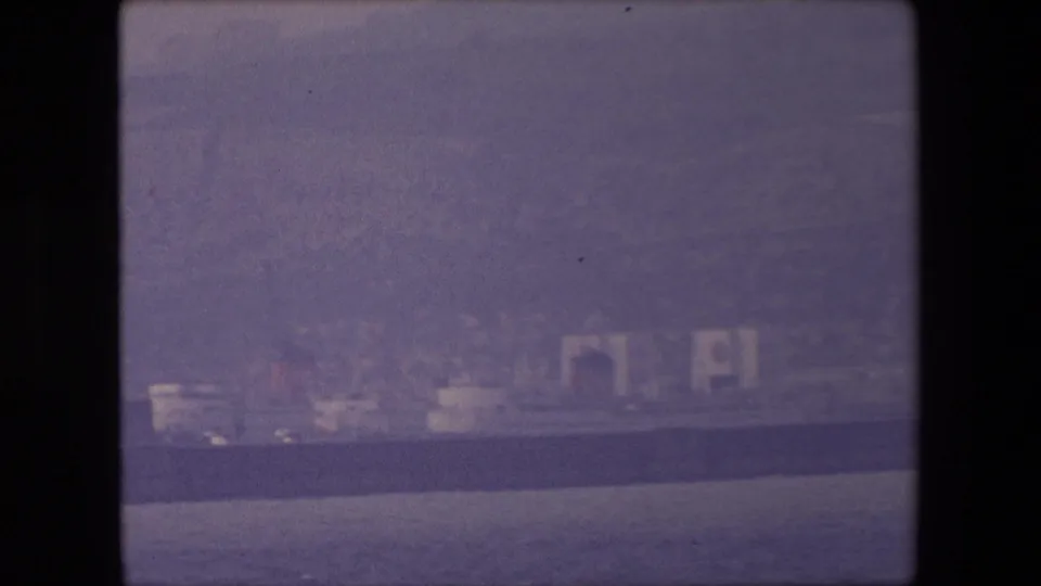 1967 8mm travel frame showing a hazy coastal view from a boat, with distant pale cliffs, dark choppy water, and soft purple color fading.