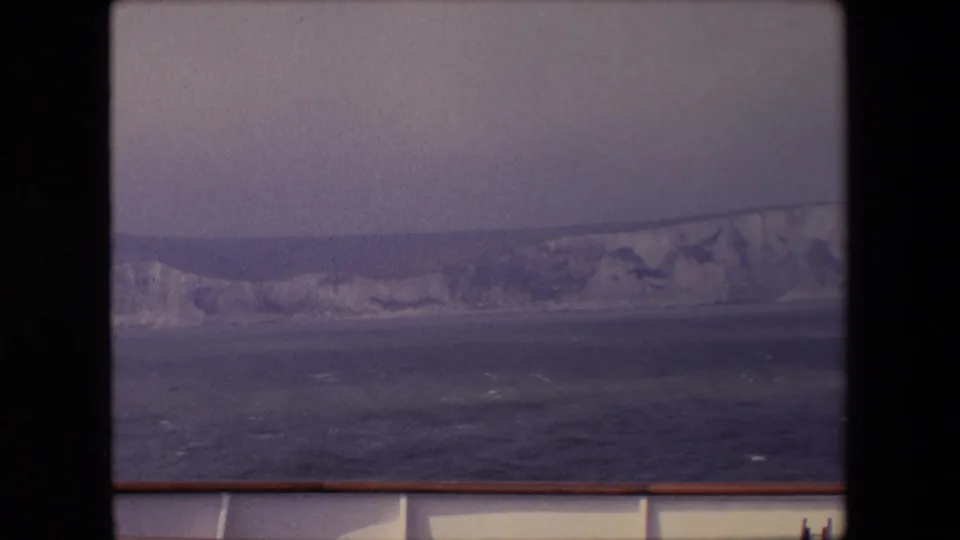 1967 8mm frame from a moving boat, showing a long pale cliff line, rough open water, muted sky, and a foreground railing.