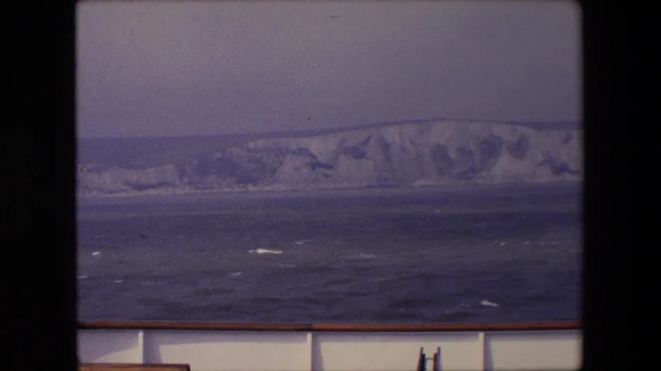 1967 England coastal travel frame with white cliffs across the horizon, dark uneven water, and the boat deck or rail crossing the lower edge.