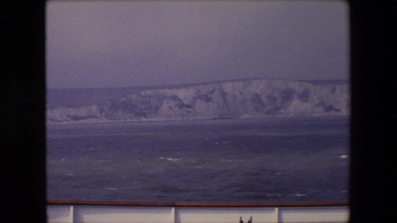 1967 8mm travel frame showing pale coastal cliffs across open water, viewed from behind a ship railing during a likely English Channel crossing.