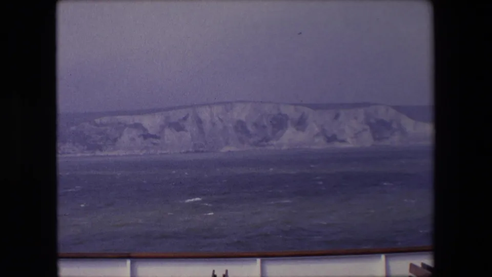 Passenger-view frame of a pale cliff coastline beyond dark blue water, with the vessel rail cutting across the lower foreground.