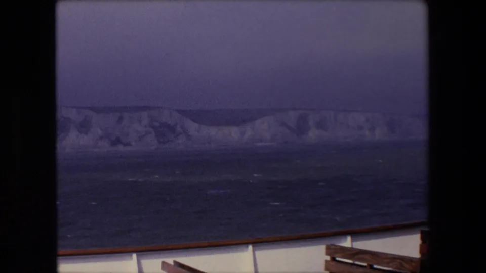 Underexposed 8mm frame showing chalk cliffs beyond dark water, with a rail or deck edge along the bottom of the frame.
