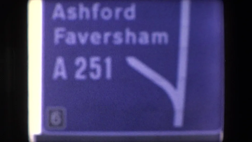 Close handheld 8mm frame of a blue directional road sign for Ashford, Faversham and the A251, likely marking the shift from coastal travel to road travel.