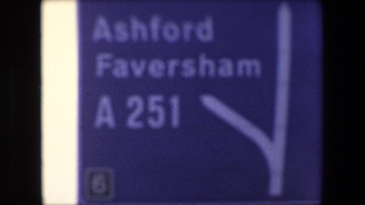 Soft close-up of a blue road sign reading Ashford, Faversham, and A251.