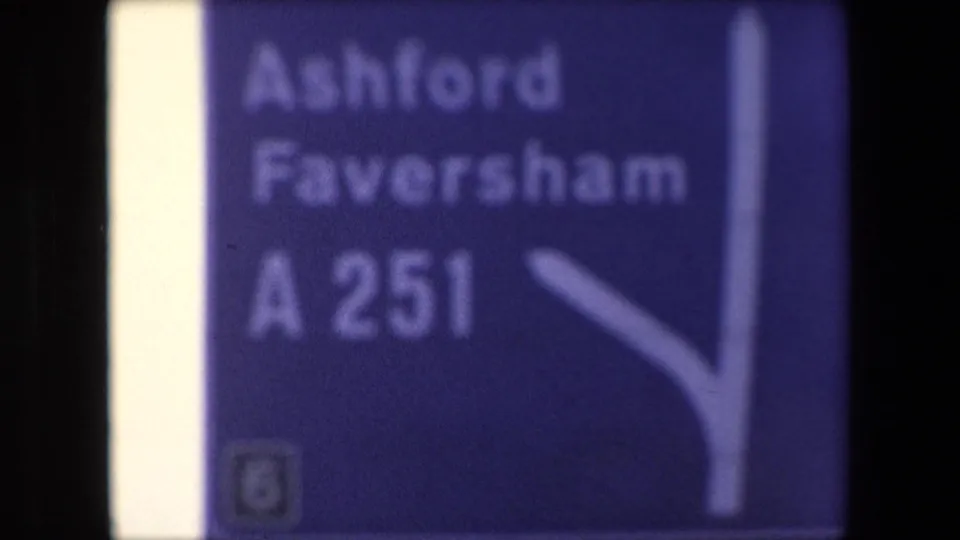 Soft 1967 London home-movie frame showing a blue British road direction sign for Ashford and Faversham via the A251, with a white arrow partly visible.