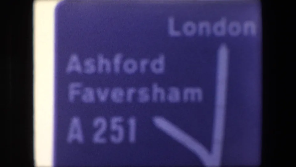 Soft 1967 London frame showing a blue directional road sign listing London, Ashford, Faversham, and A251 with a white arrow pointing upward and right.