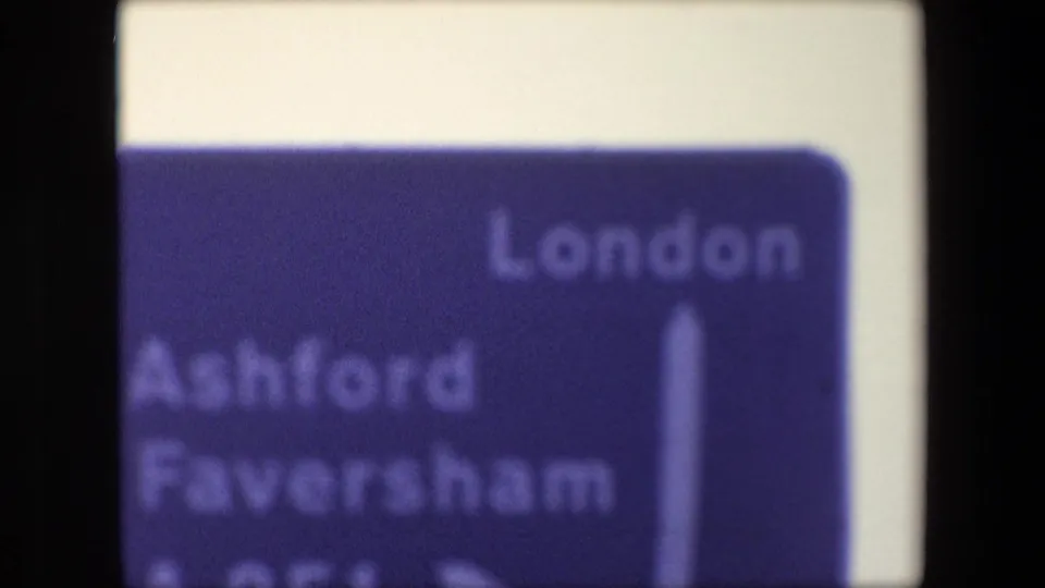 Cropped 1967 London home-movie frame showing the lower area of a blue road direction sign, with London at upper right and Ashford Faversham partly visible at left.