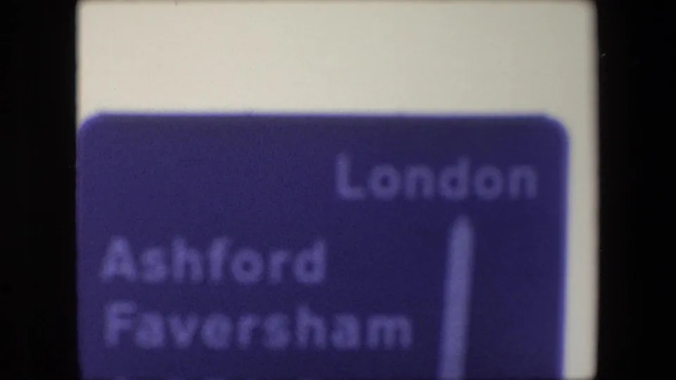 Soft 1967 London frame showing a blue road direction sign with London at the top right, Ashford and Faversham at left, and a vertical white arrow.